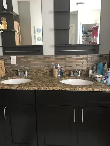 Double sink bathroom vanity with granite countertop, dark cabinets, mirror, and tiled backsplash