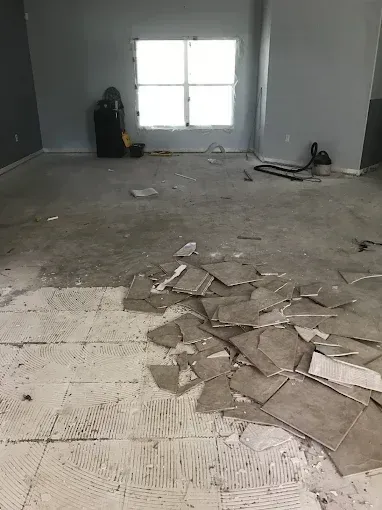 Empty room with broken floor tiles and debris near a window, suggesting renovation or demolition.