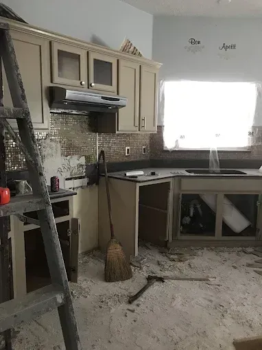 Dust-covered kitchen with cabinets, stove, sink, and debris scattered across the floor after renovation.