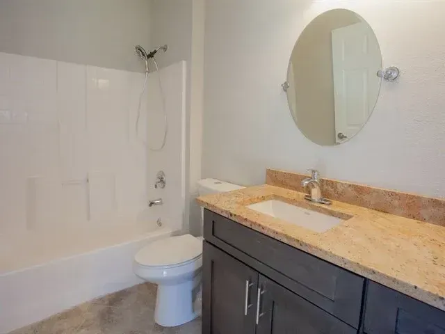 Bathroom with white tub, toilet, and dark vanity with beige countertop and round mirror.