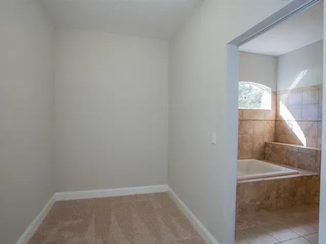 Empty beige room with carpet, open doorway, and tiled bathroom with window light