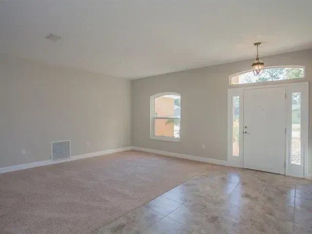 Empty beige living room with carpeted area, tiled entryway, front door, and a small arched window.