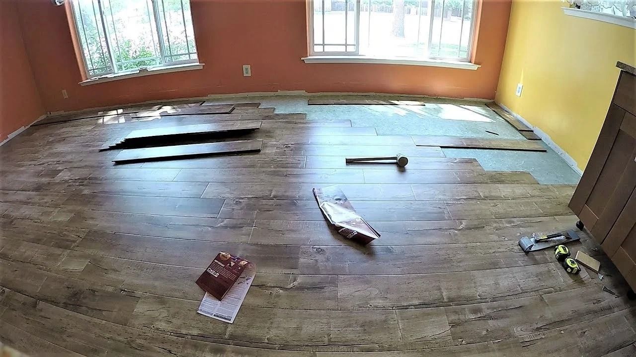 Empty room with damaged wooden floor, scattered debris, and sunlight through two windows.