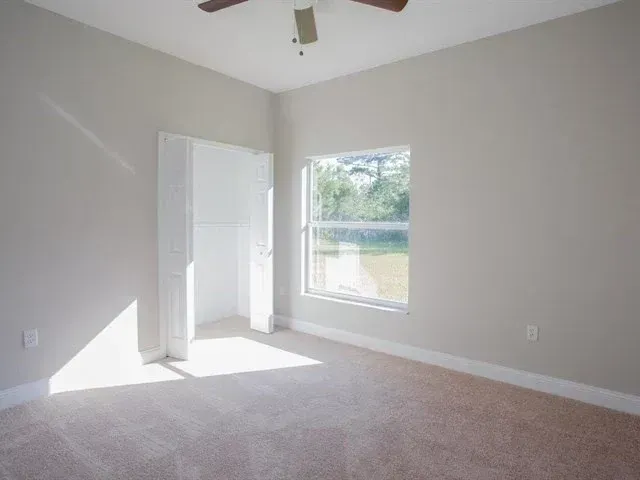 Empty beige bedroom with carpet, ceiling fan, open door, and sunlit window.