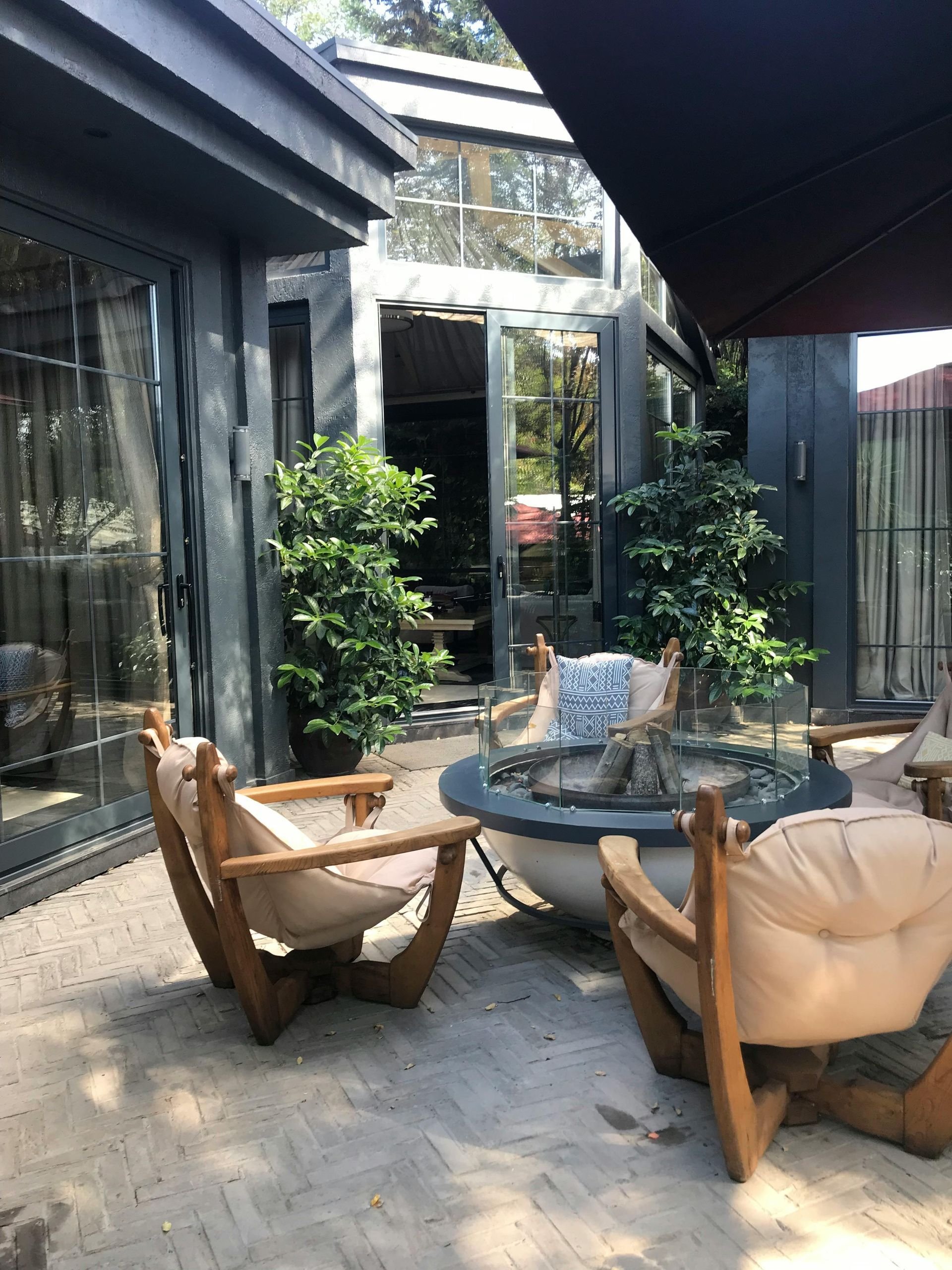 Terrasse extérieure avec sièges autour d'un foyer, à côté de portes vitrées et de verdure.