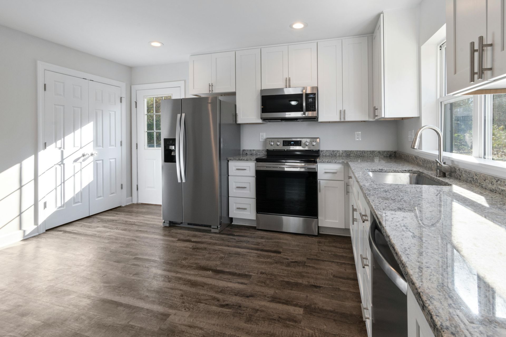 Cuisine moderne et lumineuse avec armoires blanches, électroménagers en acier inoxydable, comptoirs en granit et plancher en bois.