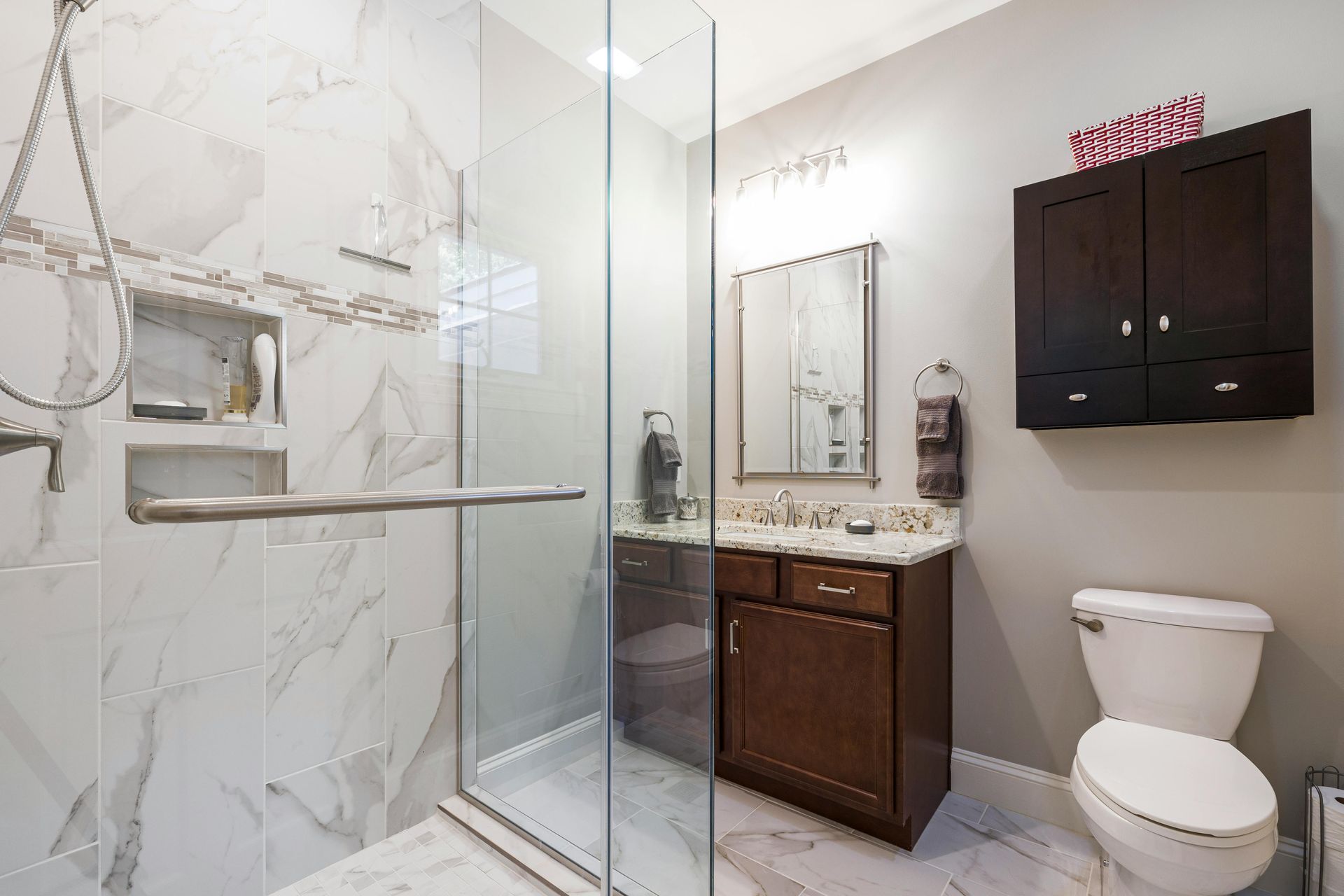 Salle de bains moderne avec douche en marbre, paroi de douche en verre, meuble-lavabo foncé, miroir et toilettes.