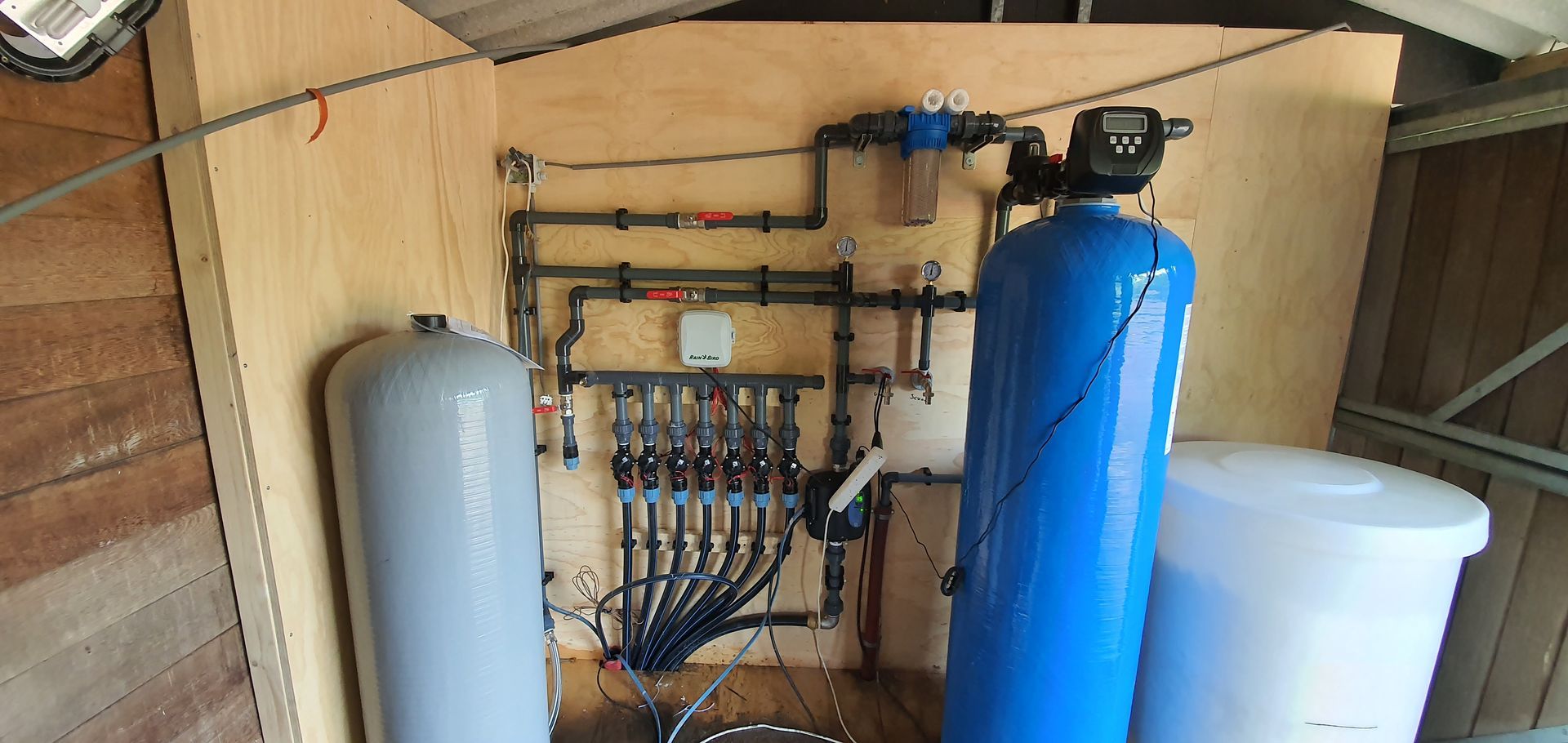 Waterfiltratiesysteem met tanks, leidingen en bedieningspaneel tegen een houten wand in een afgesloten ruimte.