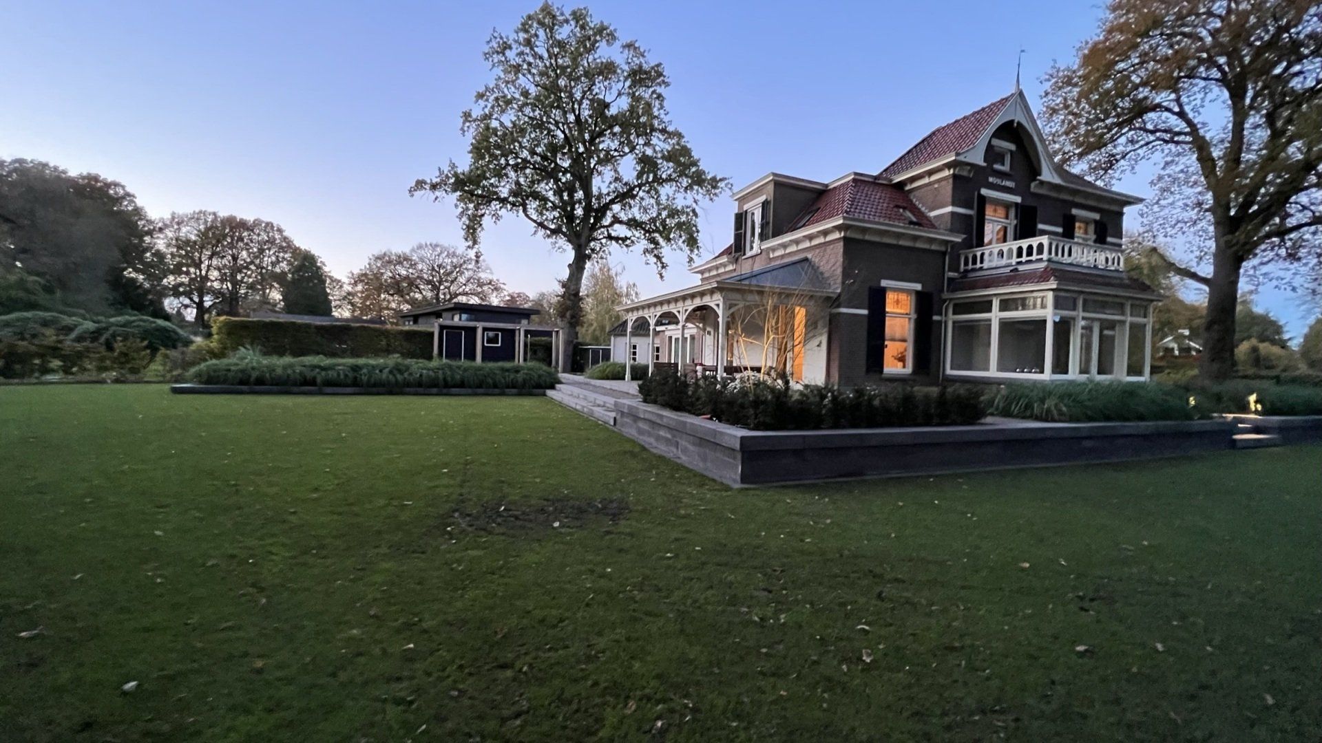 Een elegant, donker huis met een rood dak, een groot gazon en omringende bomen onder een schemerige hemel.