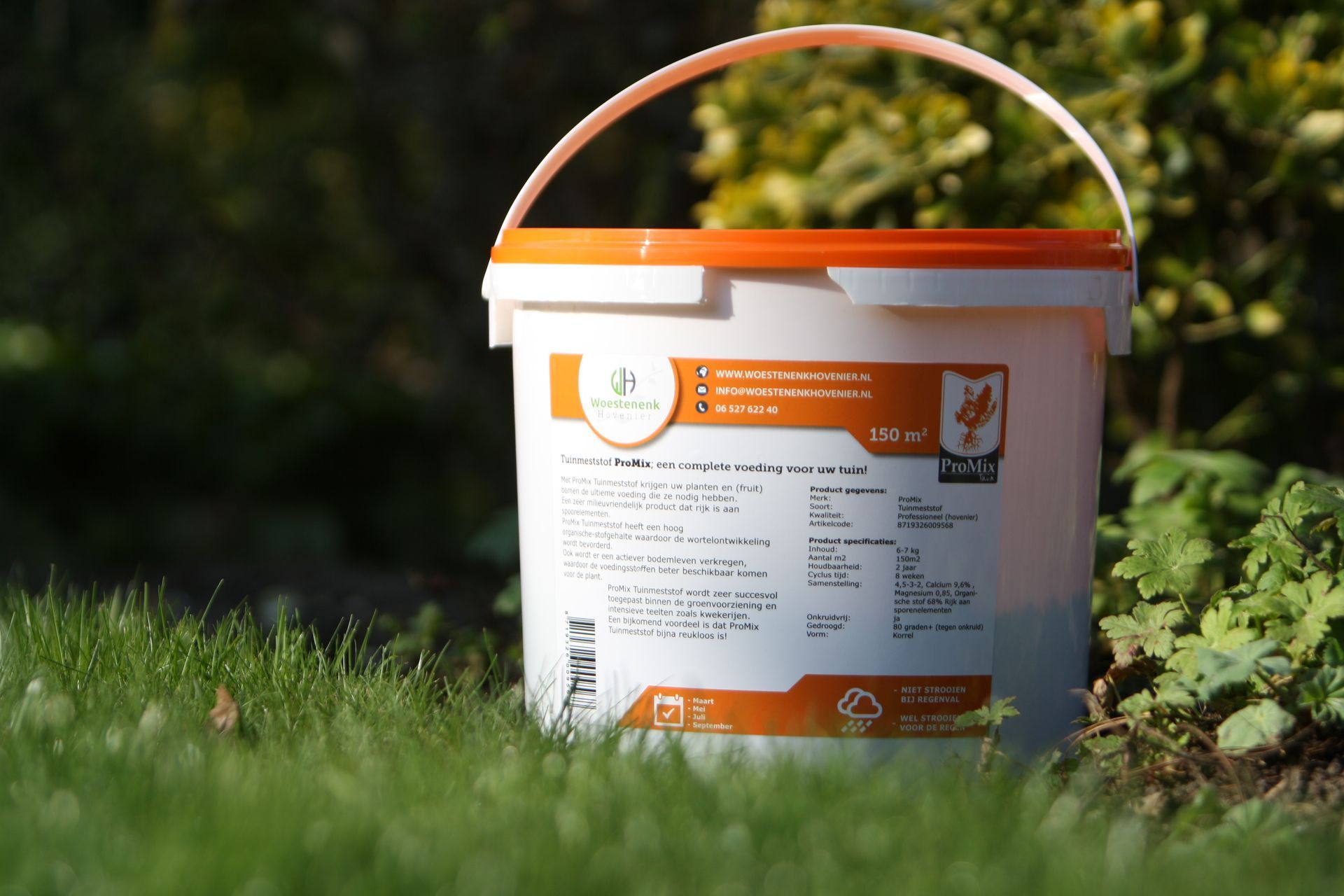 Witte emmer met oranje deksel en handvat, geplaatst in het gras, voor een groene, bladerrijke achtergrond, met labeldetails.