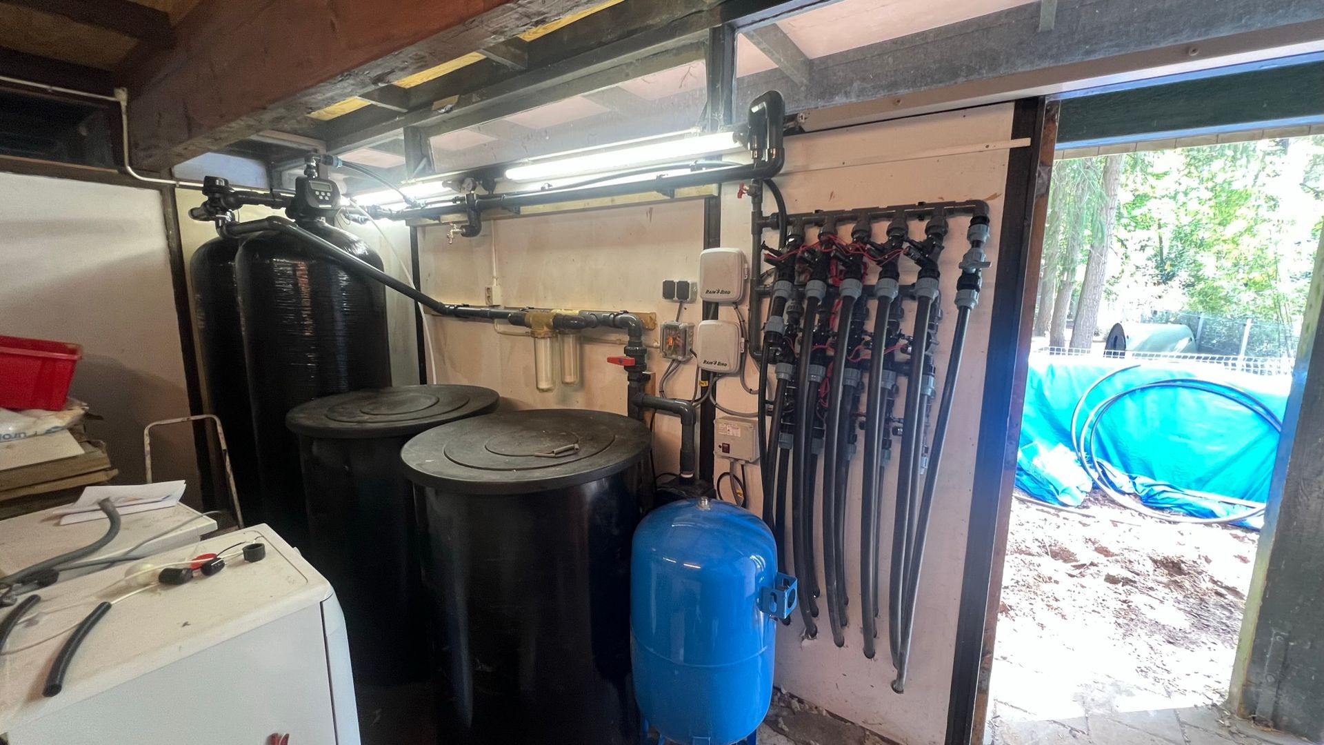 Waterzuiveringsinstallatie in een gebouw, inclusief tanks, leidingen en bedieningspaneel. Een open deur geeft toegang tot een bosrijk gebied.