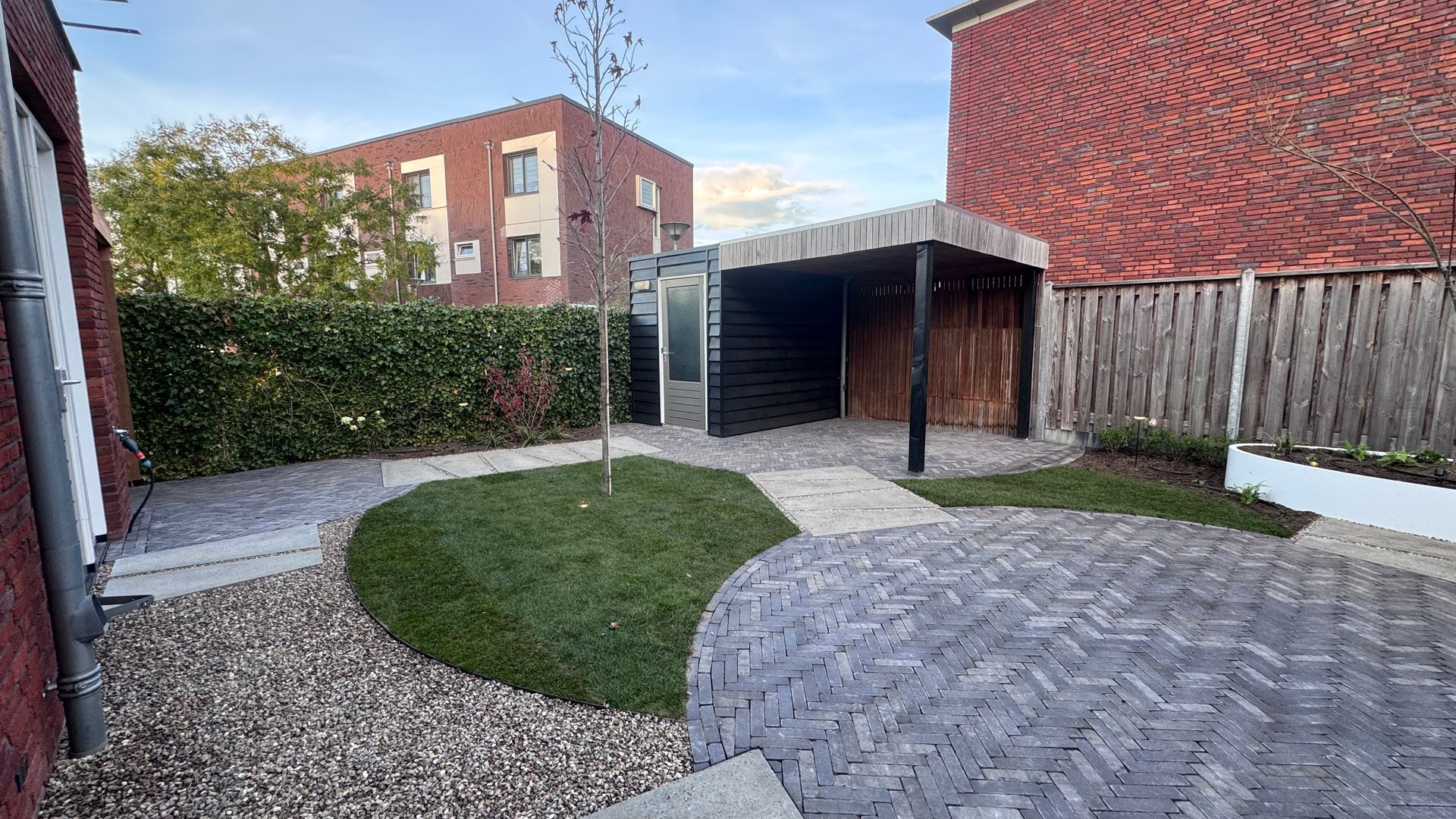 Achtertuin met grindpaden, een stenen terras, een groen gazon, een houten overkapping en rode bakstenen gebouwen.
