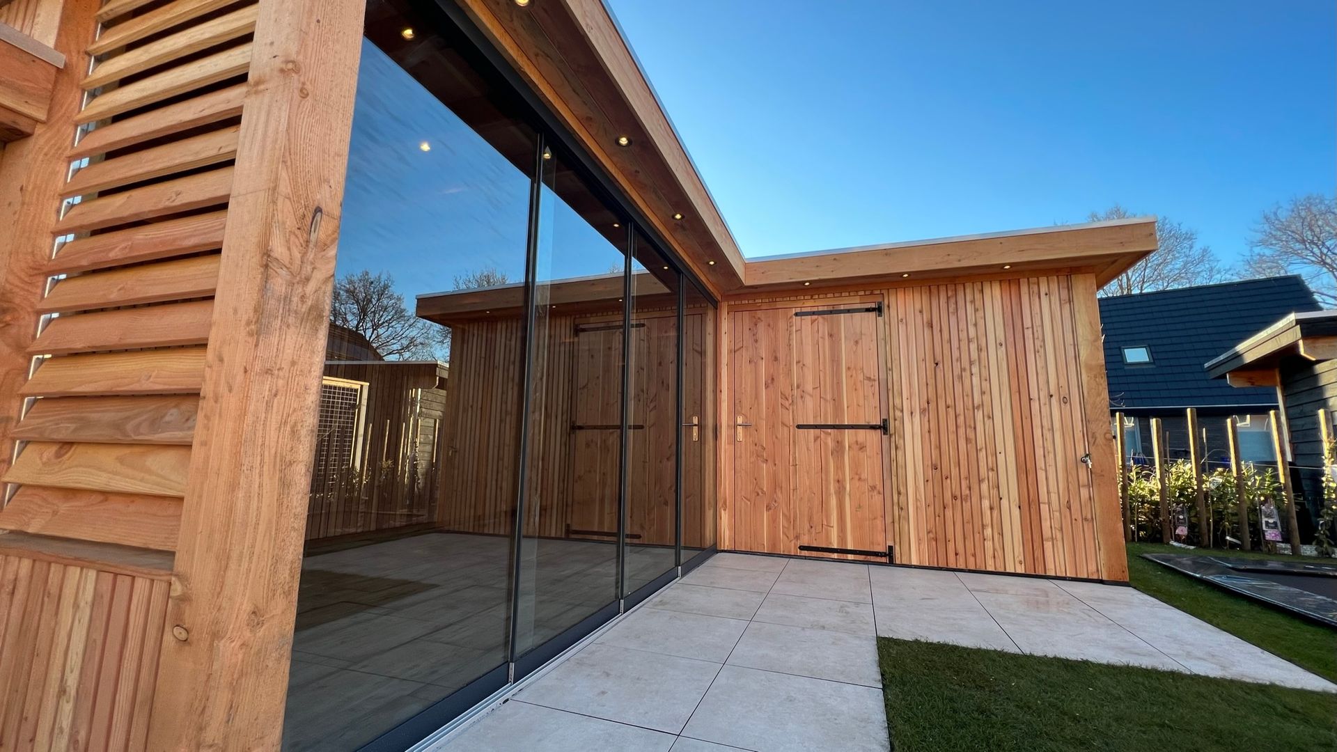 Houten bijgebouw met grote glazen ramen en deur, gelegen op een stenen terras.