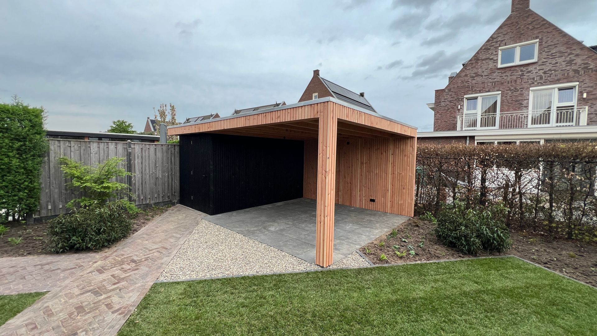 Houten carport met zwarte achterwand, grindvloer en gazon in een achtertuin van een woonhuis.