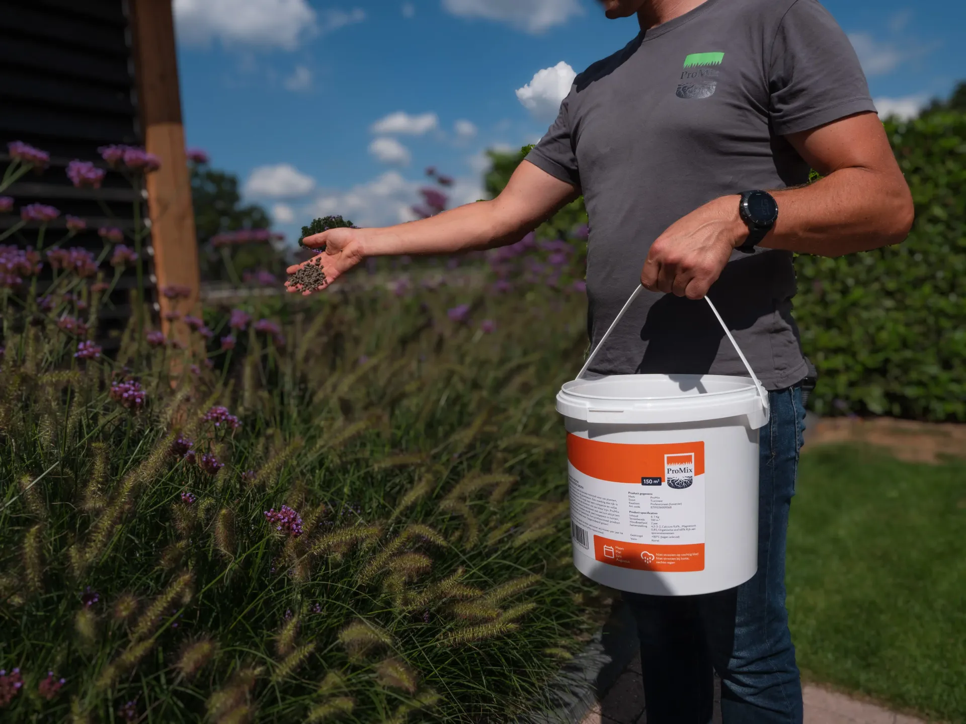 Witte emmer met oranje deksel en handvat, geplaatst in het gras, voor een groene, bladerrijke achtergrond, met labeldetails.