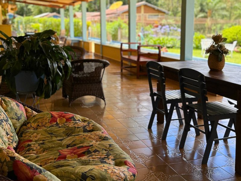 Um pátio coberto com um sofá florido, mesa de jantar, cadeiras e plantas oferece vista para um jardim e um edifício.