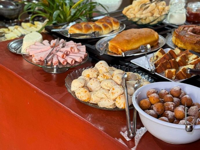 Buffet de café da manhã: pão, presunto, queijo, doces e massa frita em uma mesa vermelha.