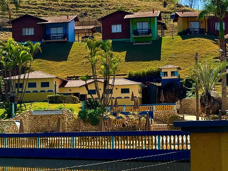 Cabanas coloridas em uma encosta, com uma cerca azul em primeiro plano e uma paisagem montanhosa ao fundo.