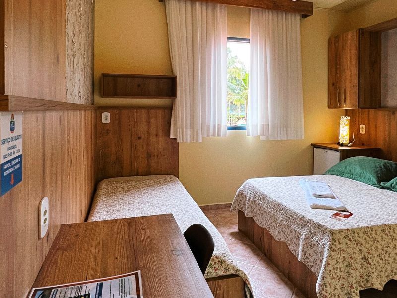 Quarto de hotel pequeno com duas camas, painéis de madeira, uma pequena janela e uma escrivaninha.