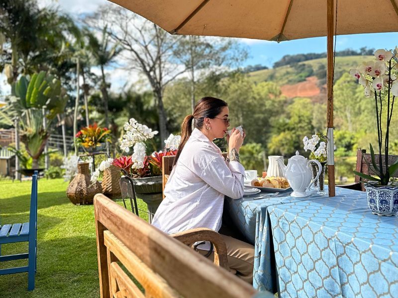 Mulher tomando chá em uma mesa ao ar livre, preparada para uma refeição. Bela vista do jardim.