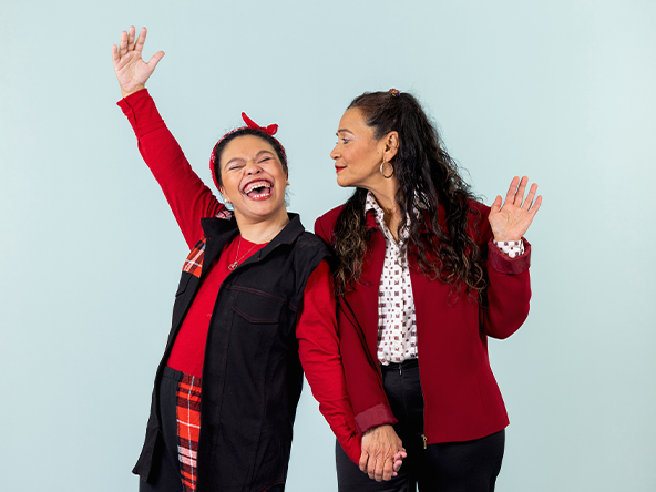 Two smiling people in red tops and black jackets wave together against a light blue background.