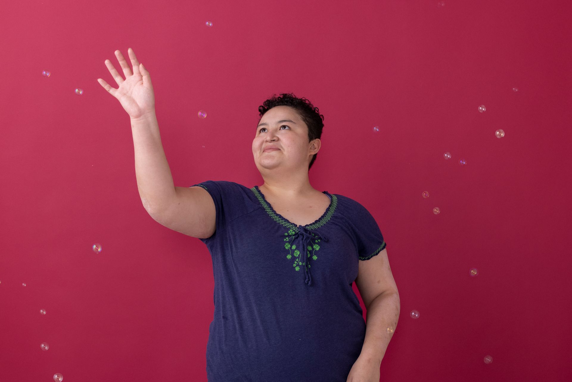 Person in a navy shirt with green embroidery raises a hand against a bright red wall.