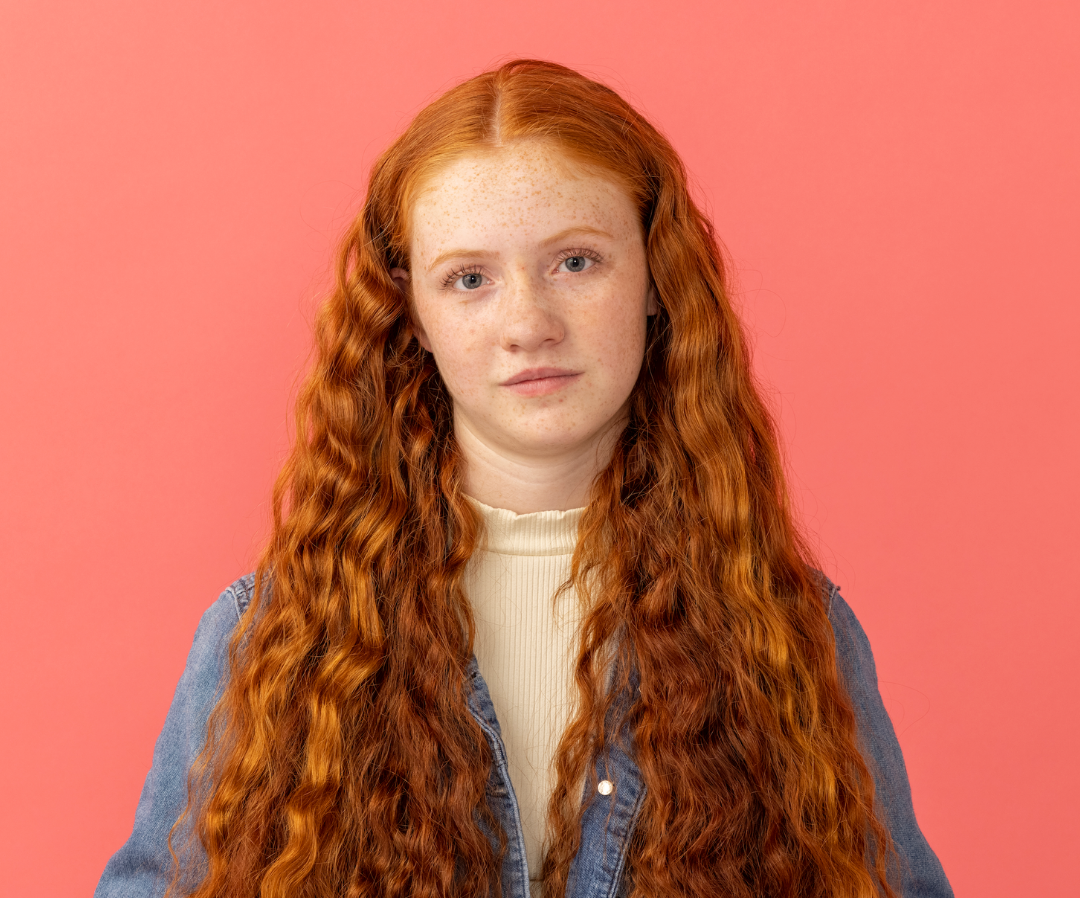 Portrait of a person with long red curly hair against a pink background, wearing a blue jacket and cream top