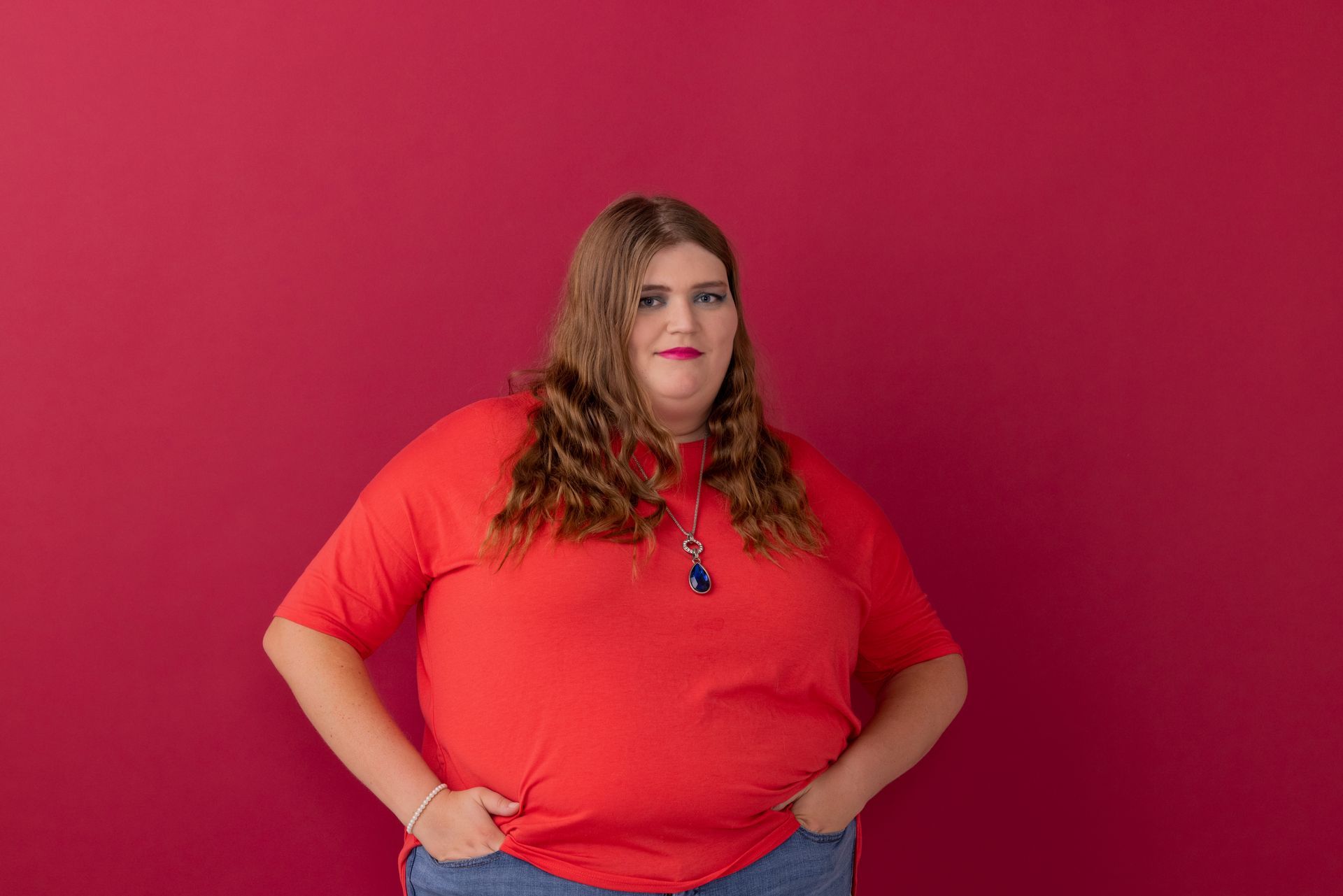 Person in a bright red shirt posing against a pink background, hands on hips.