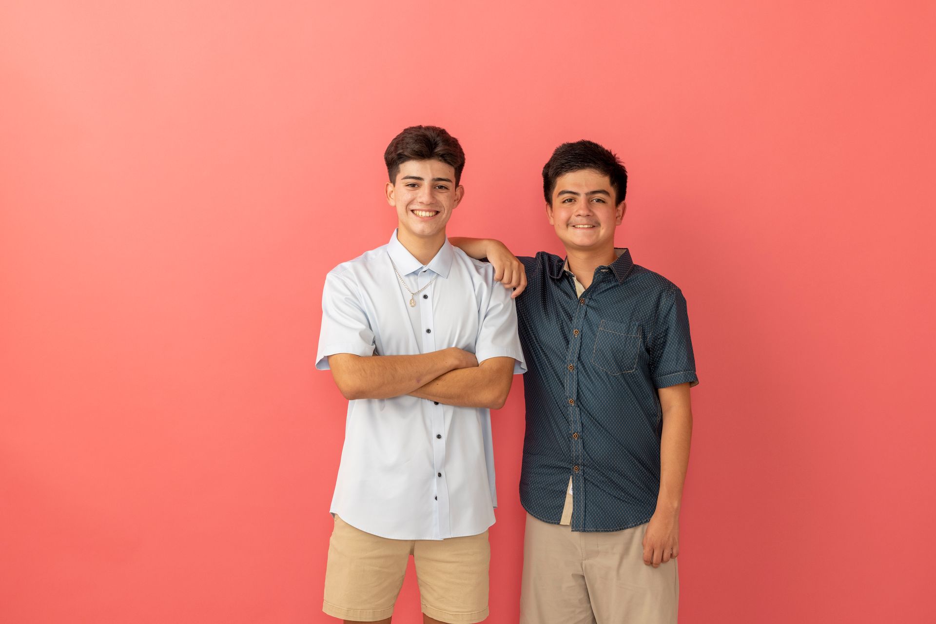 Two smiling people standing against a pink gradient backdrop, one with an arm around the other.