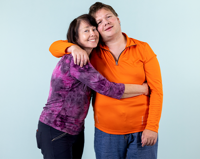Two people in orange and purple tops hugging and smiling against a light blue background