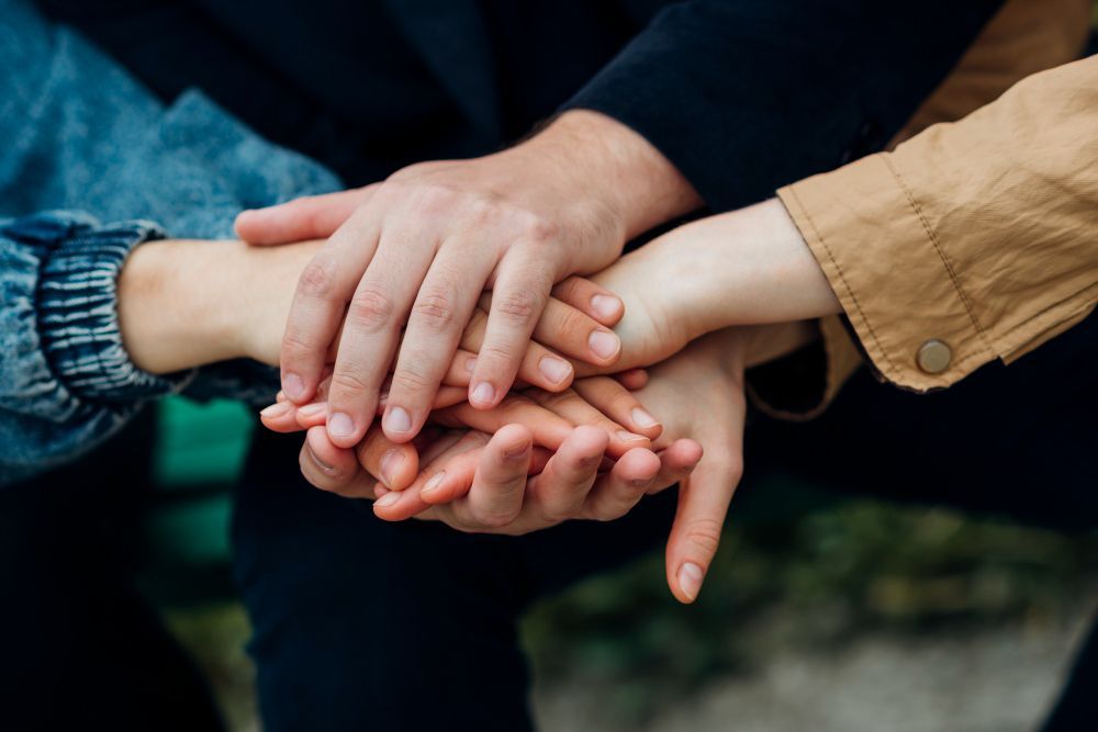 Stacked hands of four people showing teamwork, with blue and tan sleeves outdoors.
