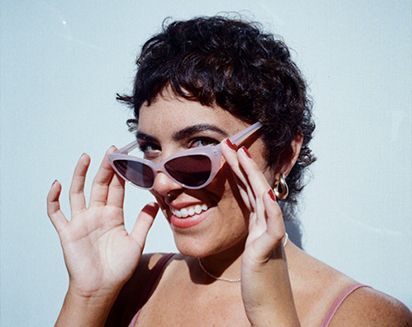 Smiling person with short curly hair adjusting white sunglasses against a light blue background