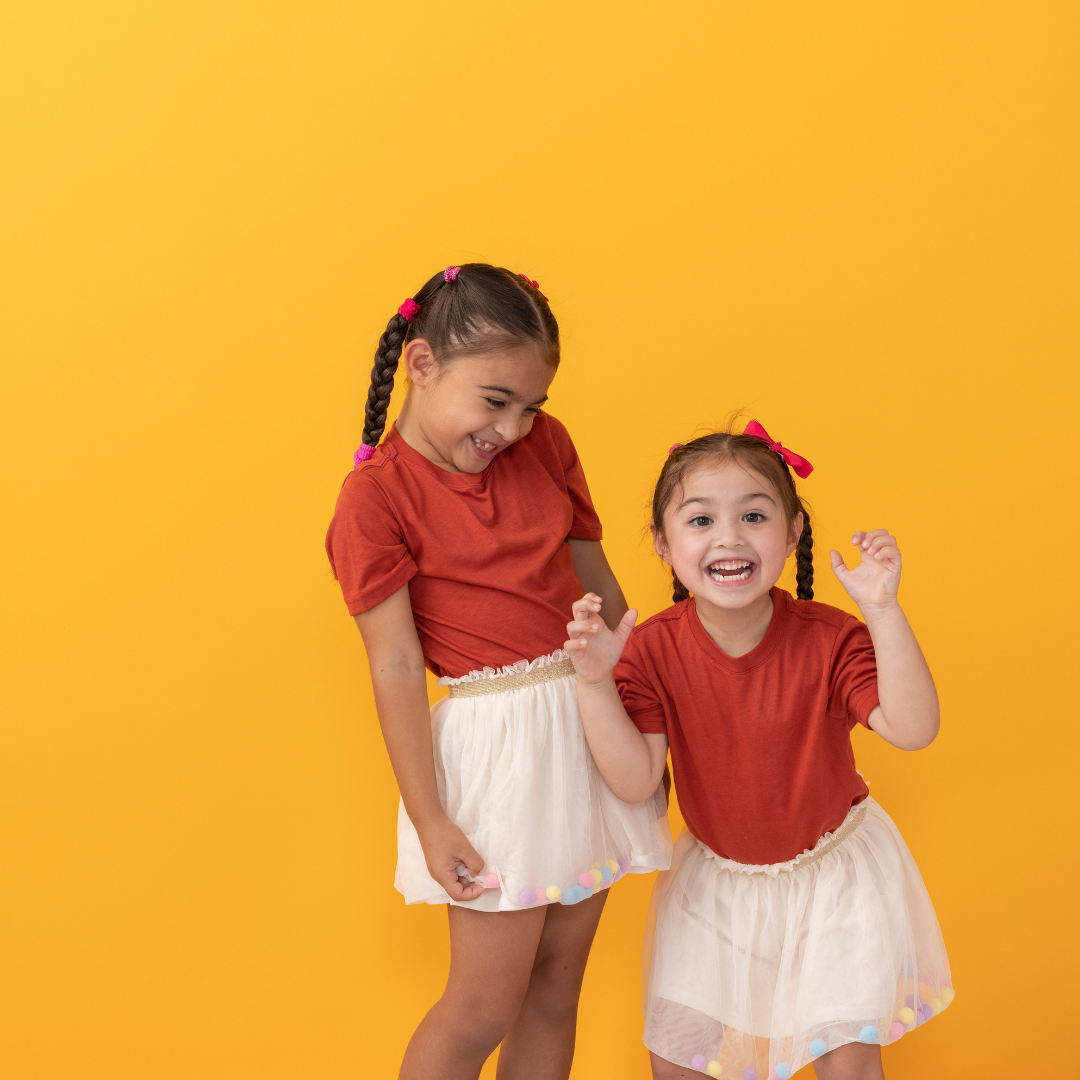 Two smiling children in red tops and light skirts posing against a bright yellow background