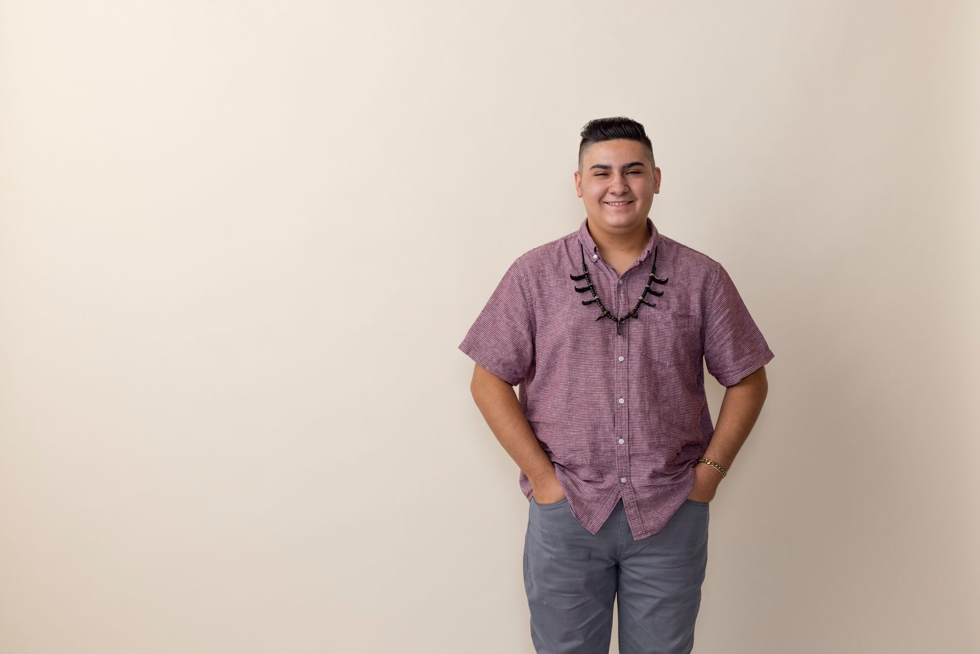 Smiling person in a pink checkered shirt and gray pants standing against a plain beige background
