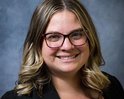Smiling person with wavy hair and glasses against a gray studio backdrop