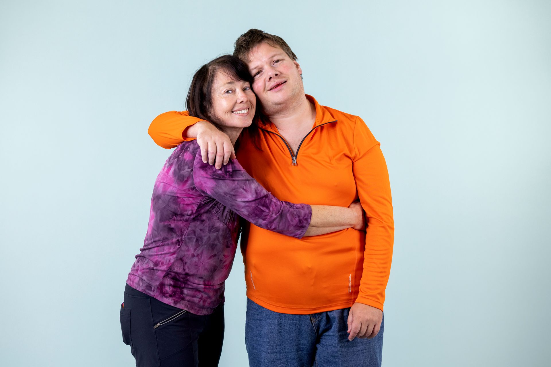 Two people hugging against a light blue background, smiling and wearing orange and purple tops.