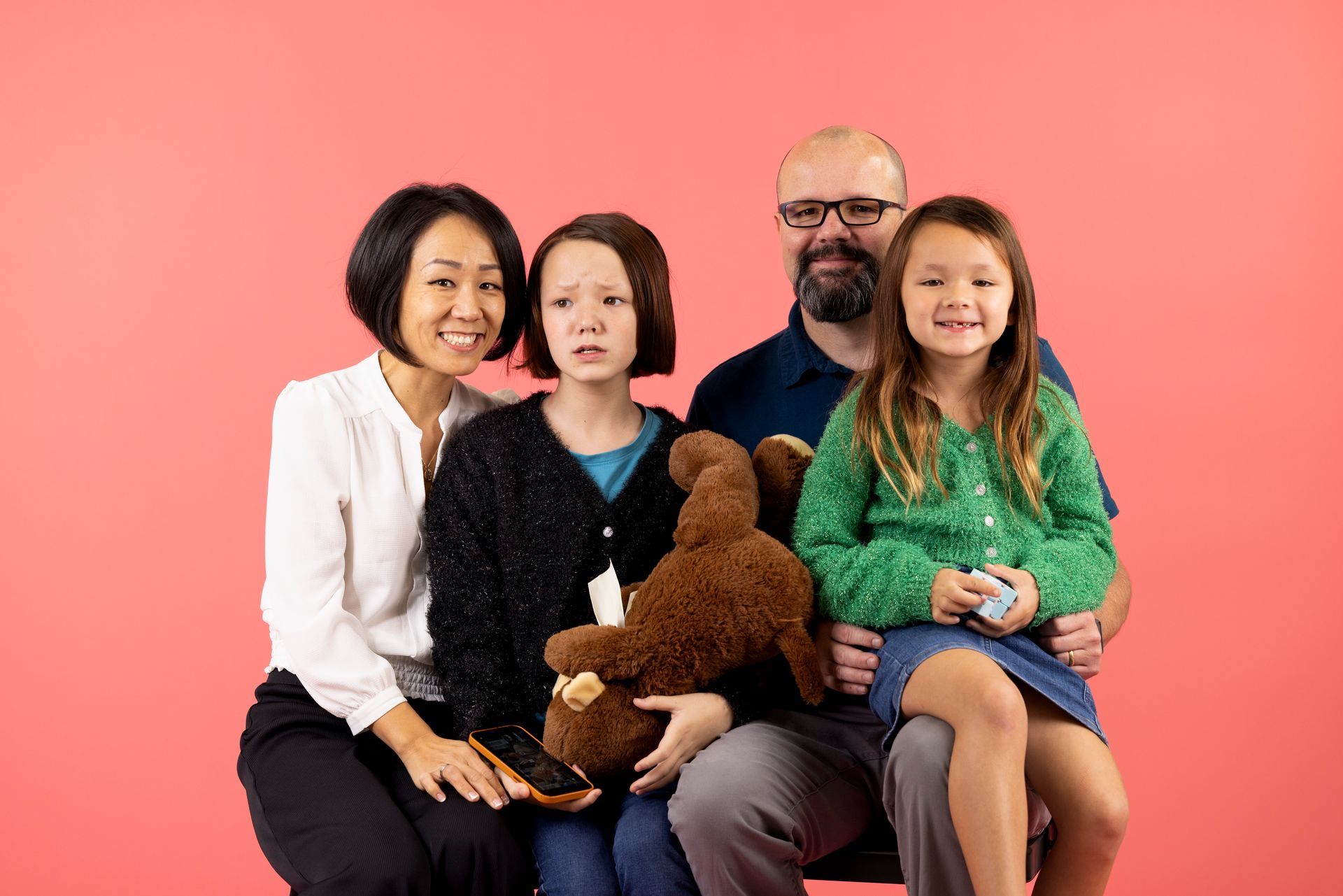Four people sit together on a couch against a pink background, holding a brown teddy bear.