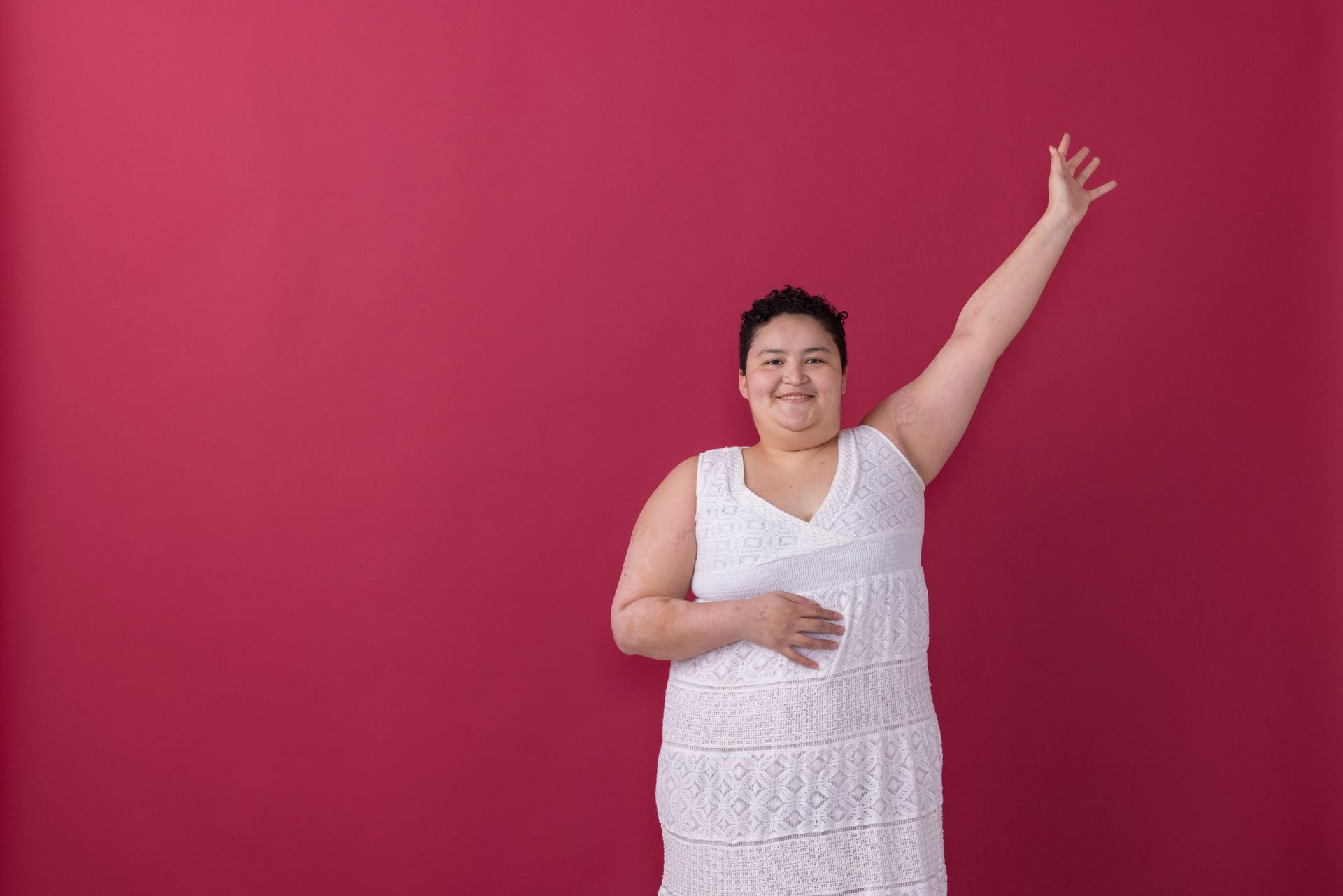 Person in a white dress smiling and raising one arm against a bright pink background