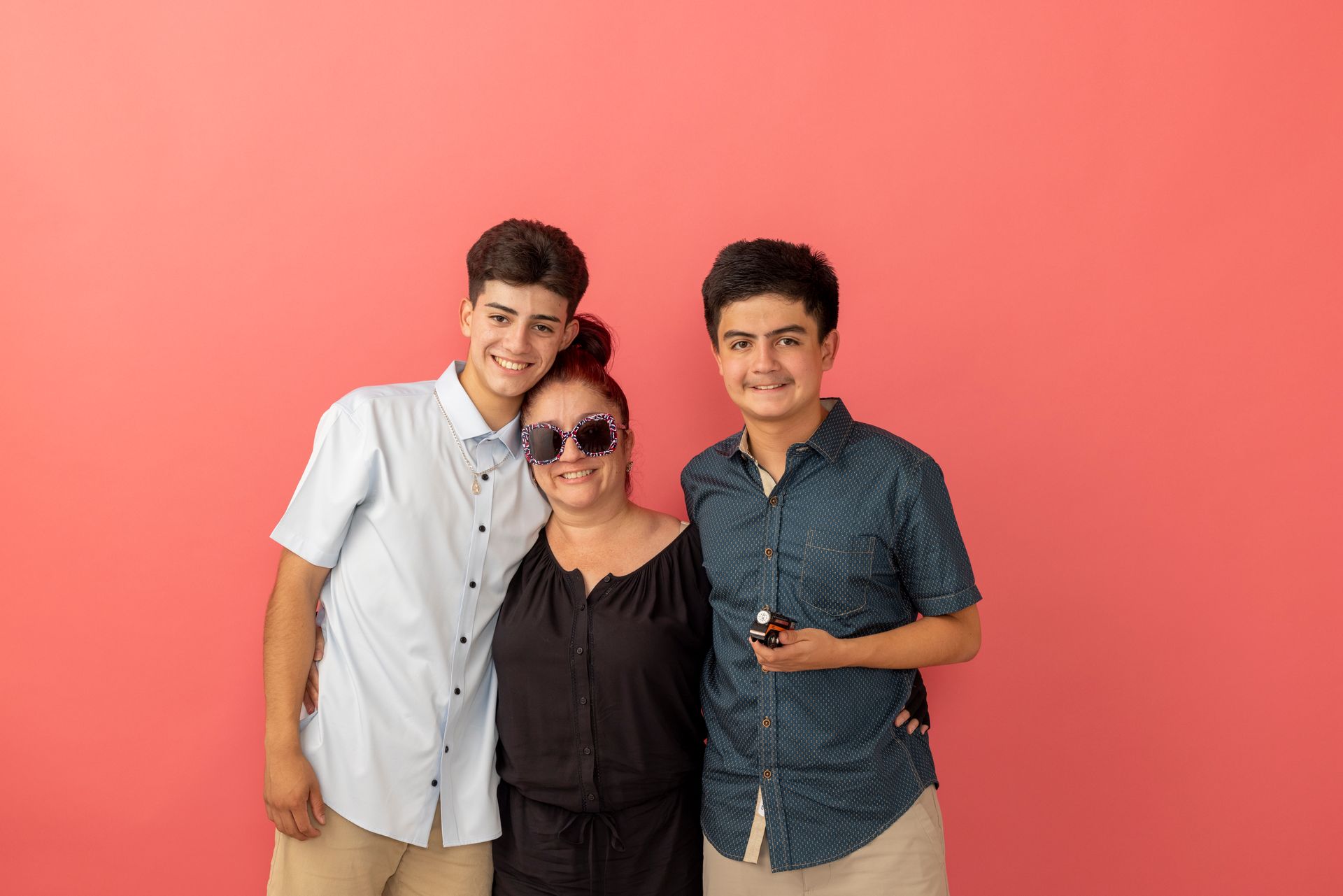 Three people posing together against a coral pink backdrop, smiling at the camera.