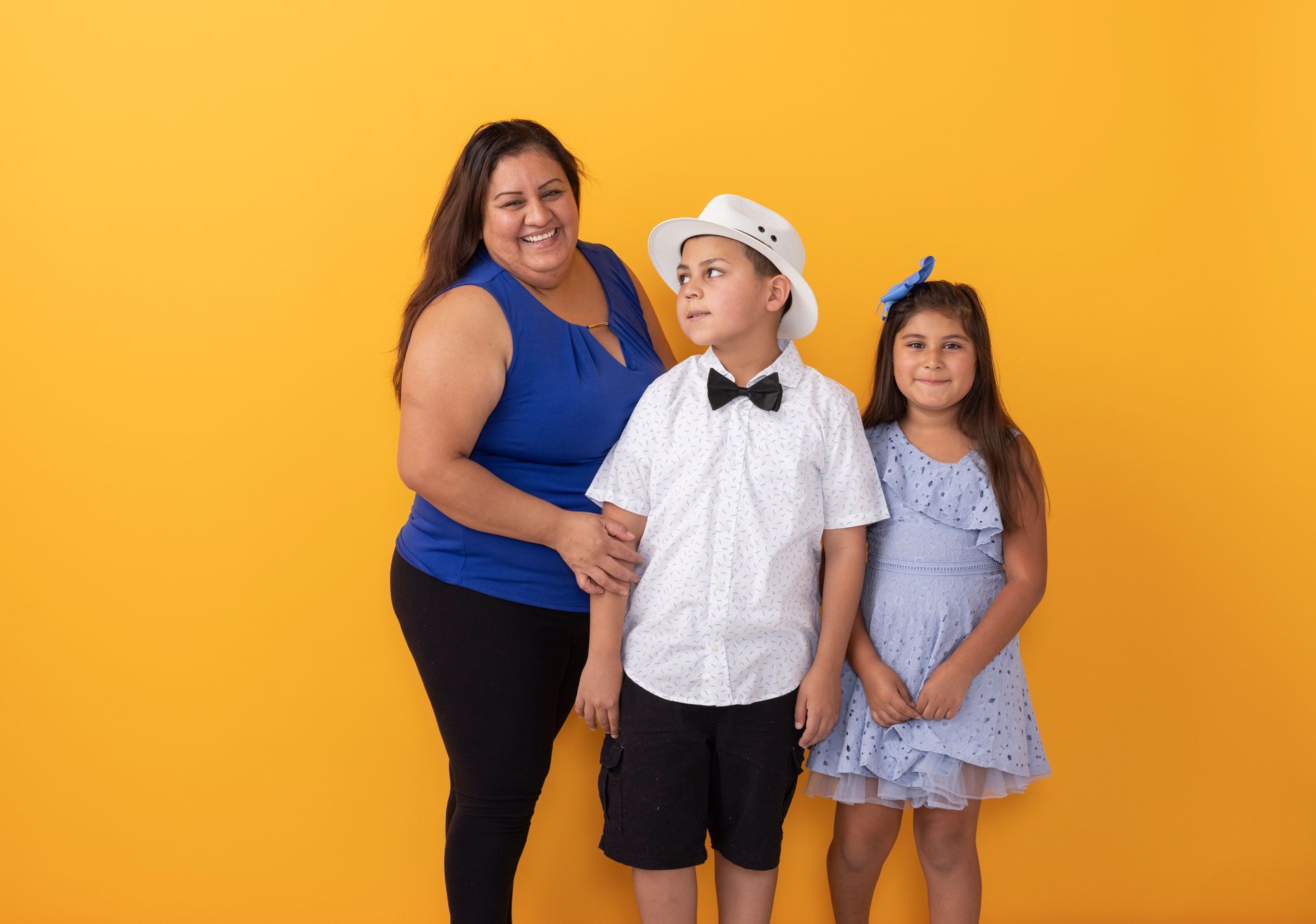 Three people posing against a bright yellow backdrop, one wearing a white hat and bow tie.
