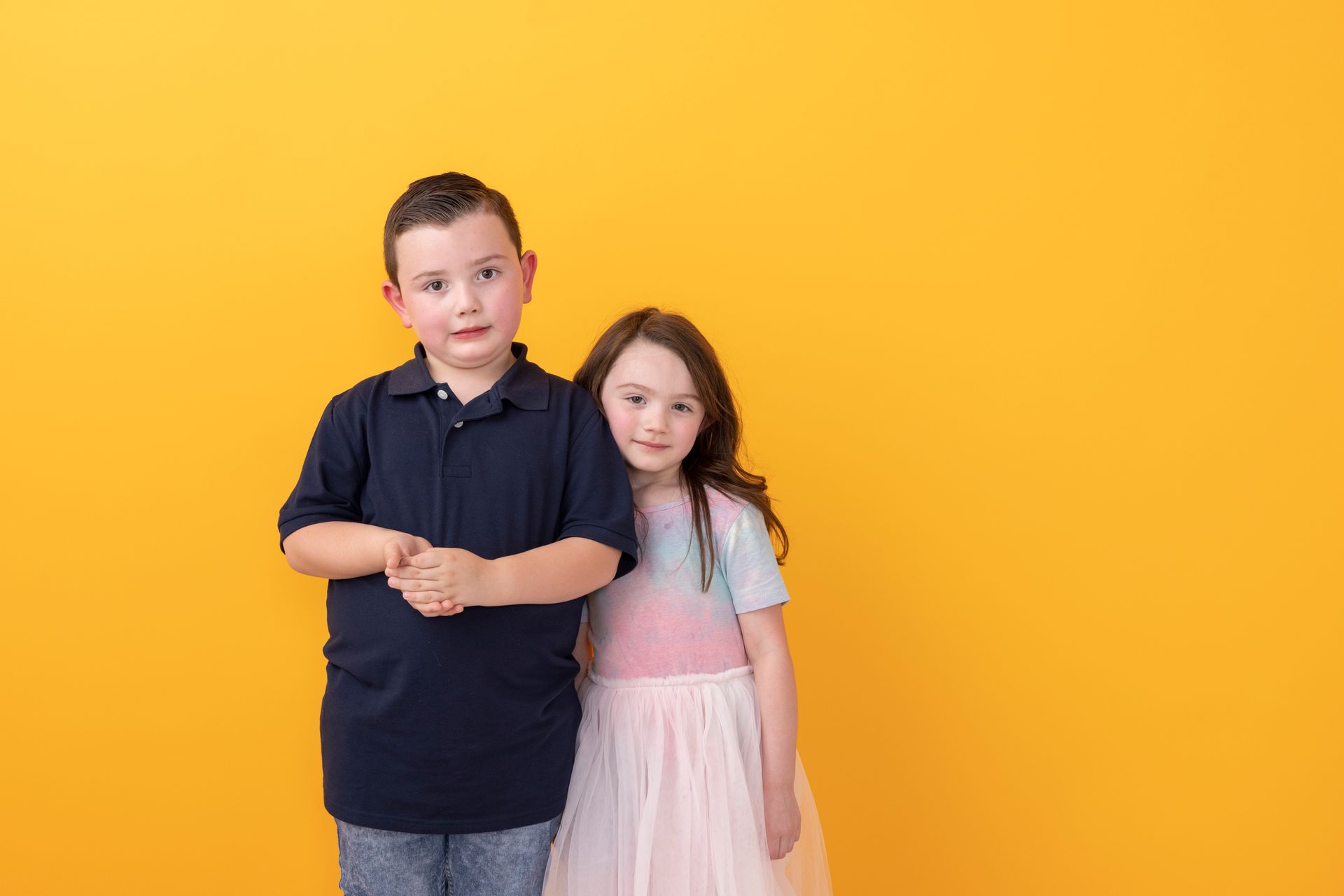 Two children standing together against a bright orange background, one in a navy shirt and one in a pink dress
