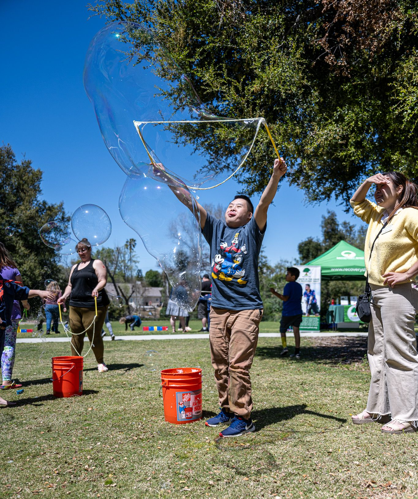 Child blowing bubbles outdoors at a park, reaching up with a wand near red buckets under a blue sky
