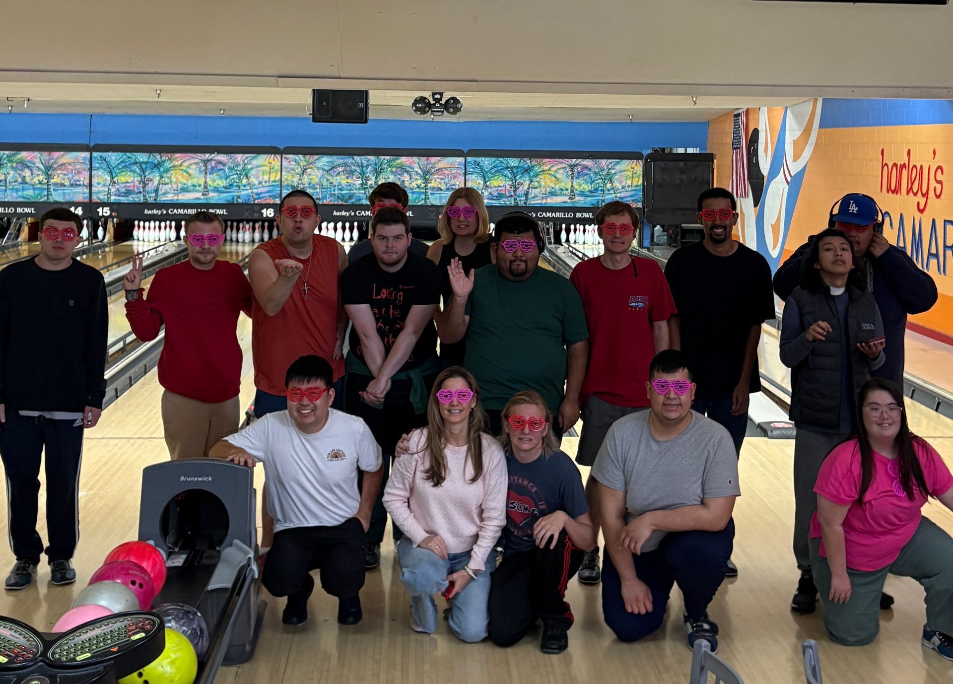 Group of people posing in a bowling alley, smiling with bowling balls and pins in the foreground.
