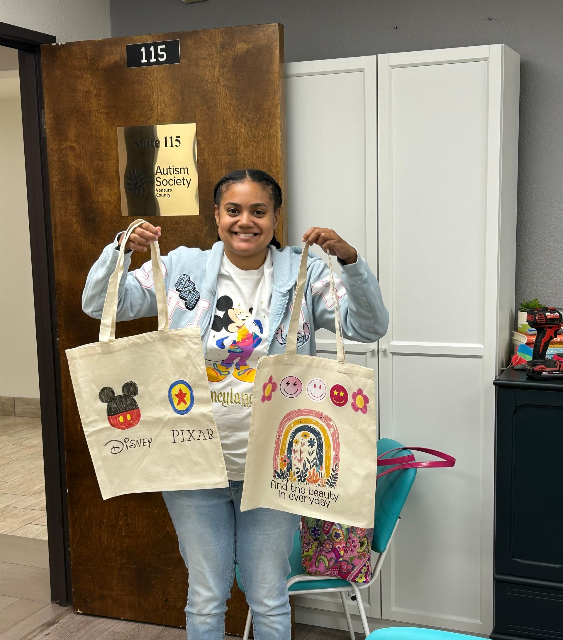 Woman smiling and holding up two decorated tote bags indoors near a wooden door and white cabinet