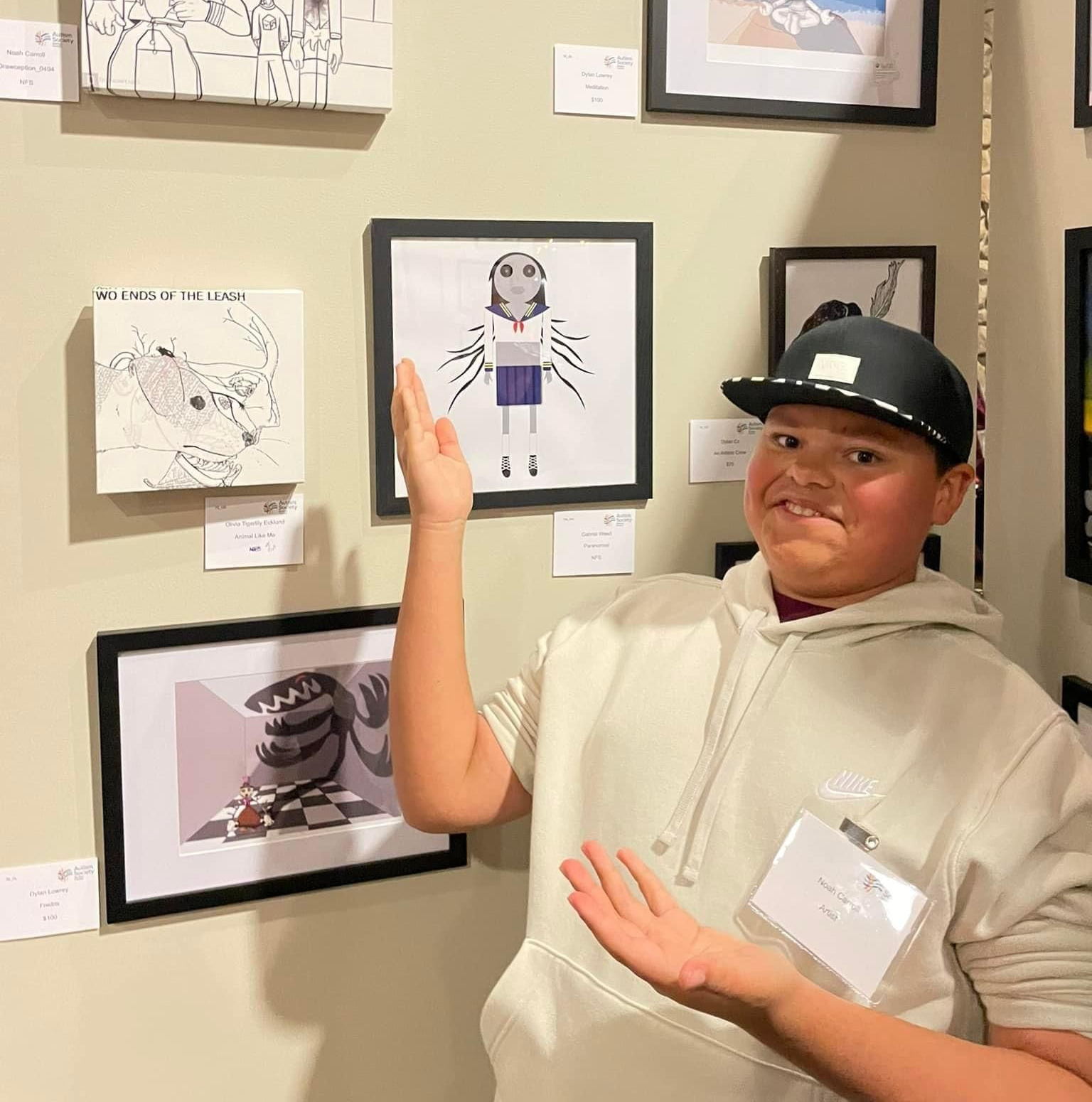 Person in a black cap and beige hoodie presenting artwork on a gallery wall.
