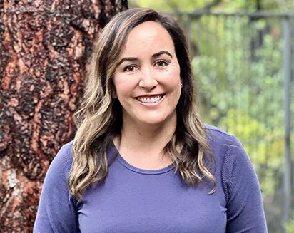 Smiling person in a purple shirt standing by a tree outdoors.
