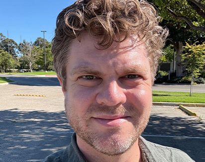Close-up selfie of a smiling person outdoors in sunny park-like surroundings