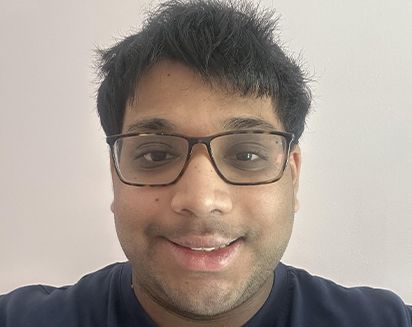 Smiling person wearing glasses and a dark shirt, facing the camera against a plain light background.