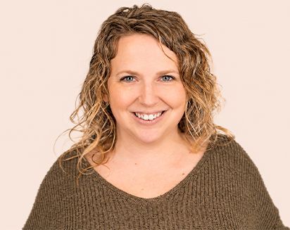 Smiling person with curly hair wearing a brown sweater against a light background