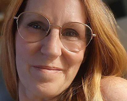 Close-up of a woman with round glasses and red hair, softly smiling outdoors in warm sunlight